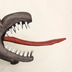 Na foto, vemos a boca de um animal esculpida em madeira, ocupando todo o lado esquerdo da foto. A boca é marrom e está bem aberta e os dentes brancos são afiados. De dentro da boca, sai uma lingua vermelha fina, que ocupa todo o centro da foto. O fundo da foto é bege.