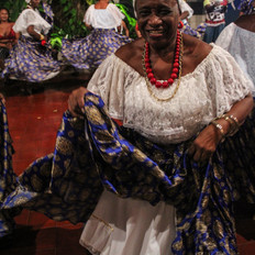 In the photo we see a woman in Creole drum clothes: white dress, turban and a round flower skirt. She moves her skirt while she smiles. In the background we see other women in wheel dancing in the same clothes.