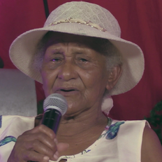 In the photo, we see an elderly, black woman wearing a hat. We only see her from the bust up. In the background, we see a pink cloth and a part of a grey percussion instrument. She holds a microphone.