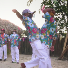 Na foto vemos dois homens batendo suas coxas, numa menção a uma luta. Eles vestem calças brancas e camisas azuis claro com estampas floridas. O cenário da foto é um terreno de areia com árvores ao fundo.