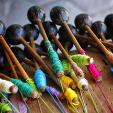 Fotografia colorida mostra em detalhes pequenas hastes de madeira, também conhecidas como bilros, utilizadas para tecer renda. A imagem mostra em torno de dezoito hastes amontoadas. Estão dependuradas numa almofada por fios amarelos, verdes, cor-de-rosa, em tons de azul e bege. Os bilros medem mais ou menos quinze centímetros e possuem na extremidade oposta aos fios, pequenos cocos de tucum encaixados, o que lhe atribui formato semelhante a um pirulito. A parte superior da fotografia, onde estão as extremidades das hastes com os cocos de tucum, aparece levemente desfocada, enquanto do meio para baixo, parte que mostra o corpo das hastes e as linhas coloridas, aparece bem nítida, de modo a destacá-las.