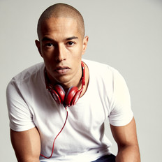 Bald brown man wearing a white shirt with short sleeves and with a red headset hanging around his neck, He's sitting, but we only see his body from the waist up. He's looking directly at the camera. The background of the photo is slightly greyish.