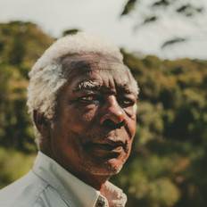 Na foto, vemos o rosto de um homem negro, idoso, cabelos brancos. Vemos a gola de sua camisa, branca. O fundo está desfocado, com vegetação verde. Ele está levemente inclinado para a esquerda, mas o rosto está virado pra a frente, olhando diretamente para a câmera.