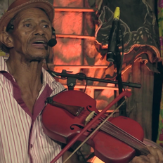 Na foto, vemos um homem negro, com microfone auricular e chapéu de couro pequeno. Ele veste uma camisa de listras vermelhas. Ele toca uma rabeca, instrumento semelhante ao violino.
